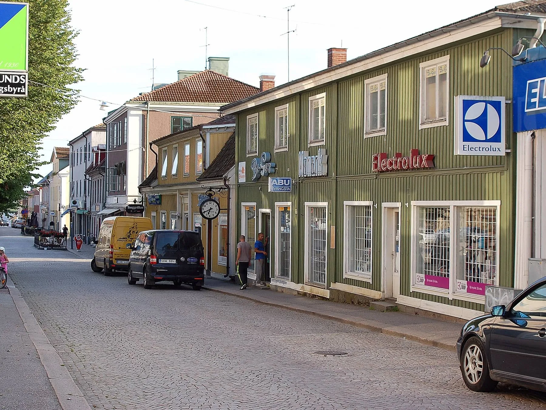 Storgatan i Askersund med gatsten, trähus i grönt och gult och butiker i bottenvåningarna.