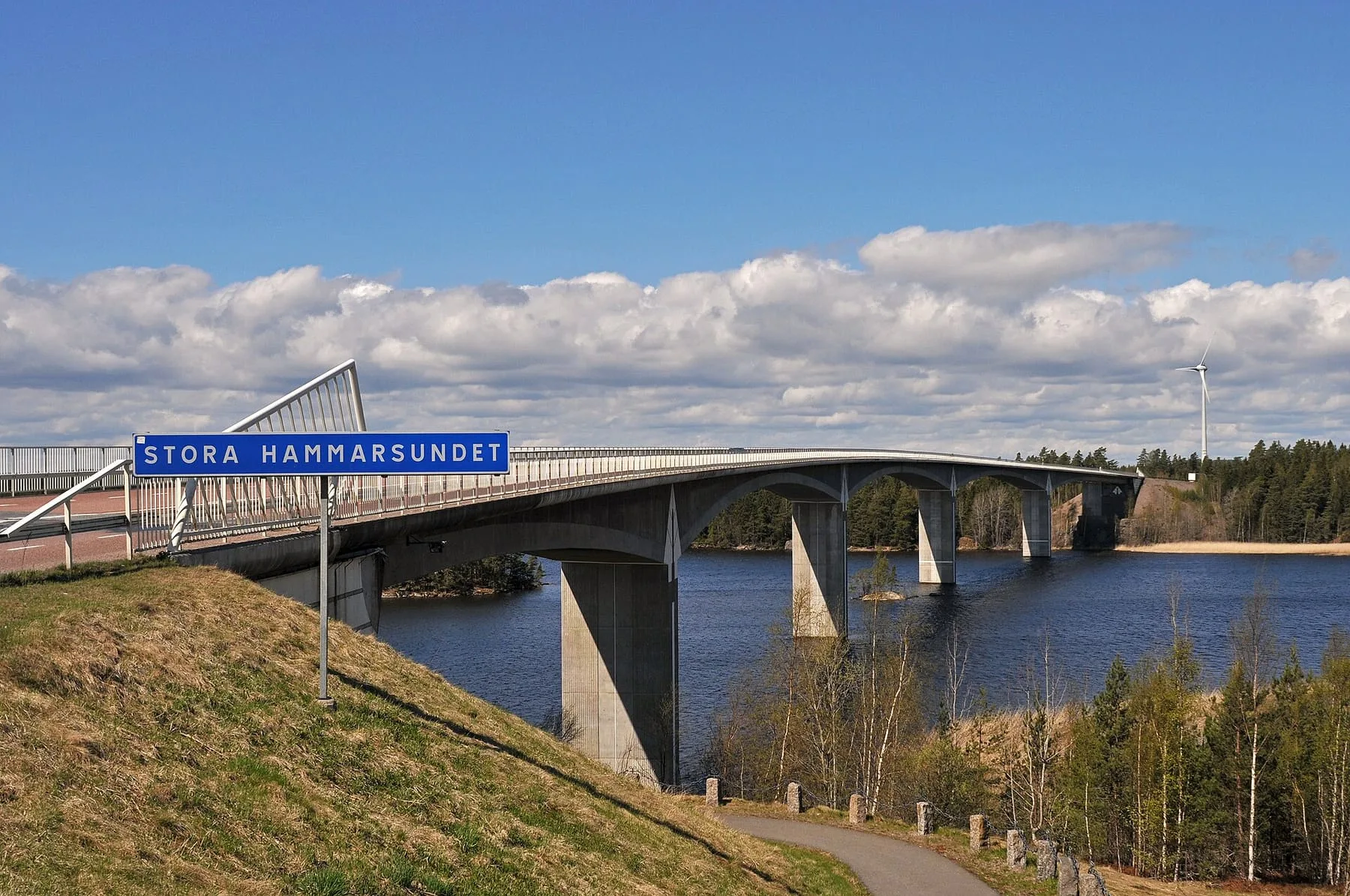 En bro över ett smalt sund med blått vatten, skog på båda sidor och en båt under bron.