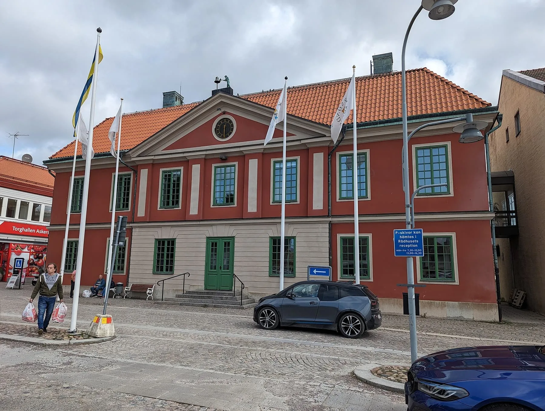 Askersunds rådhus, en röd tvåvåningsbyggnad med vita detaljer och flaggstänger framför, vid den stenlagda torgytan.