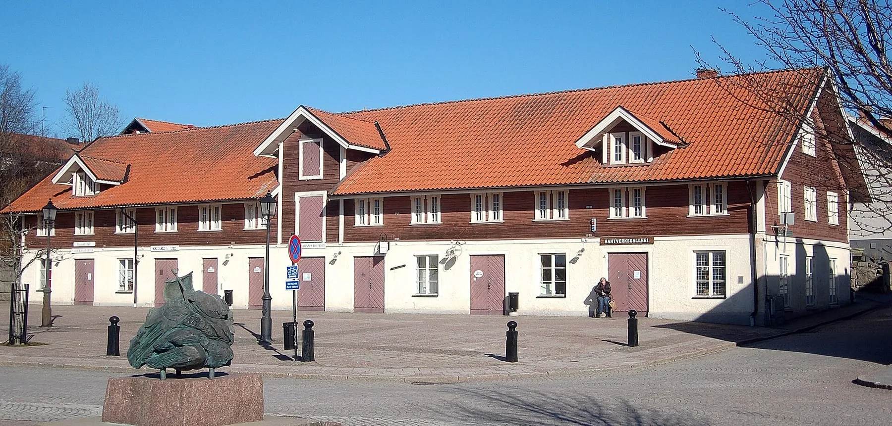 Hamnmagasinet i Askersund, en lång byggnad med rött tegeltak och portar i bottenvåningen, en skulptur på den stenlagda platsen framför.