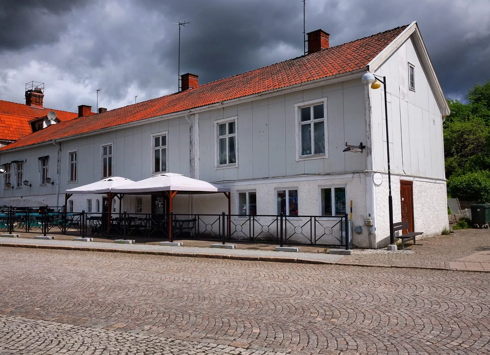 Långt vitt trähus i två våningar med rött tegeltak och uteservering under parasoller vid en gata med gatsten.
