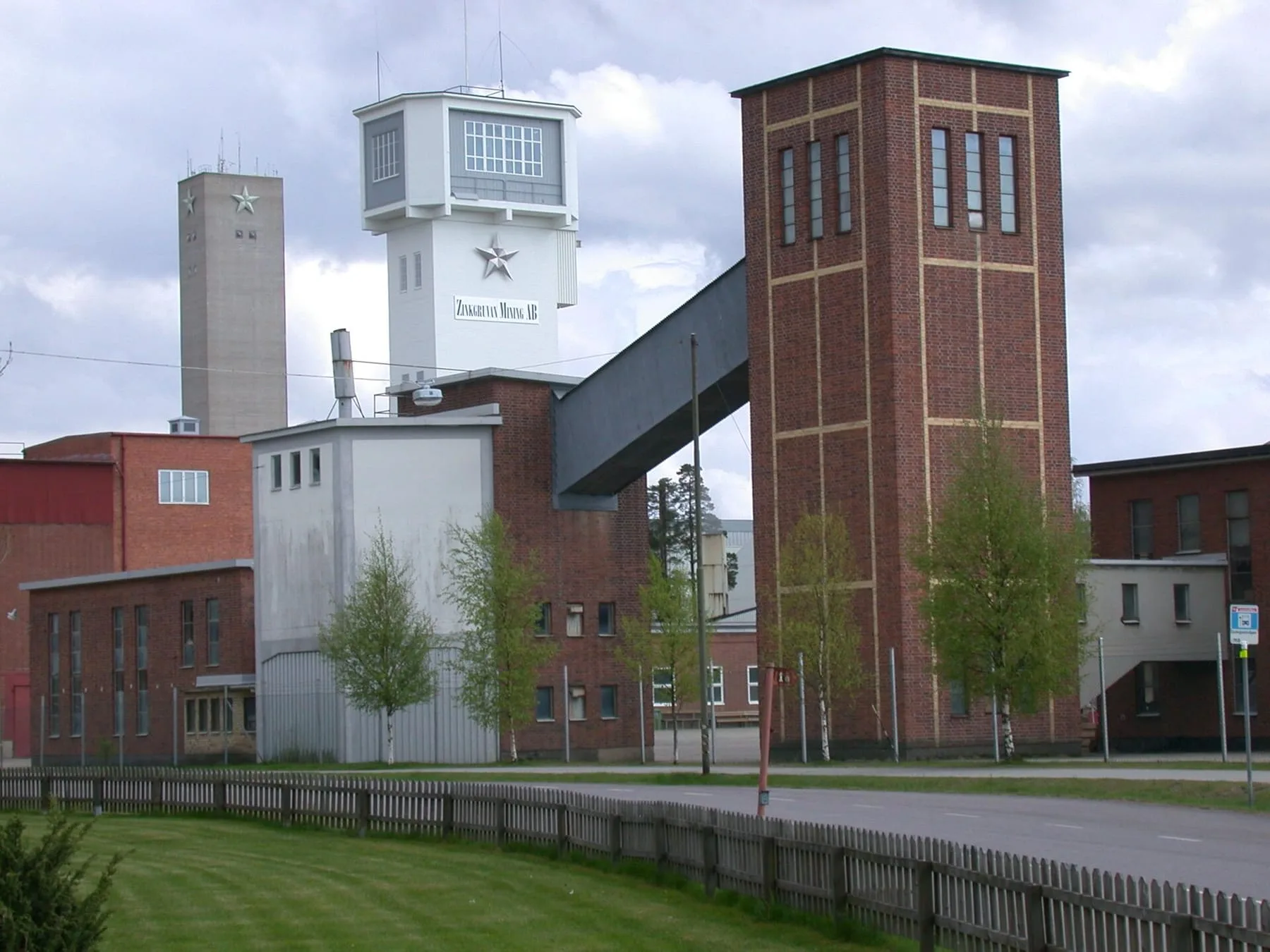 Gruvanläggningen i Zinkgruvan med ett högt tegeltorn, en vit lavbyggnad och industrihus bakom ett staket.