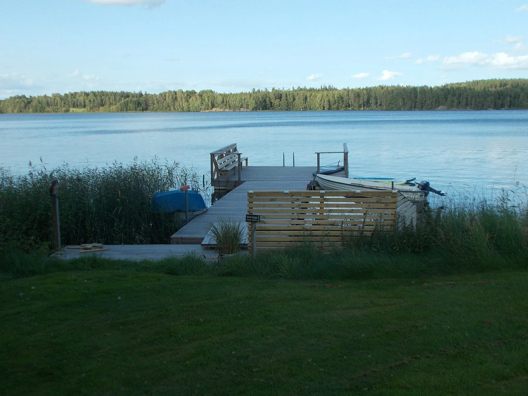 En brygga och en liten båt vid en stilla sjö med skogsklädd strand på andra sidan.