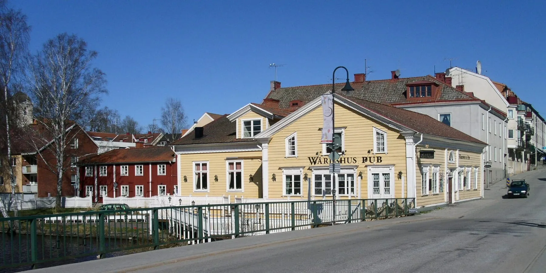 Det gula värdshuset i Askersund med skyltar för rum, värdshus och pub, vid en bro med räcke och röda trähus i bakgrunden.