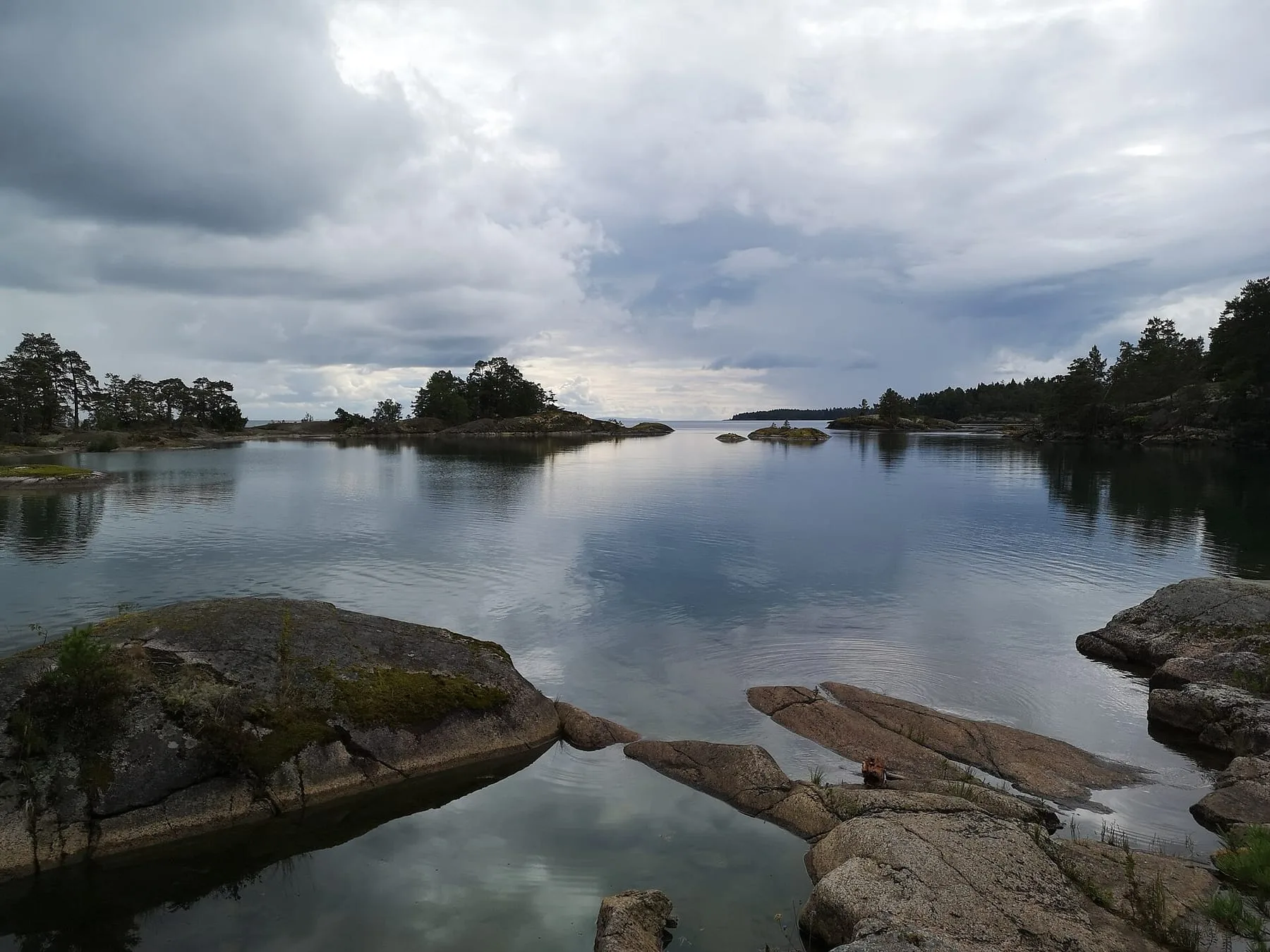Spegelblankt vatten mellan släta berghällar och små skogklädda holmar under en molnig himmel.