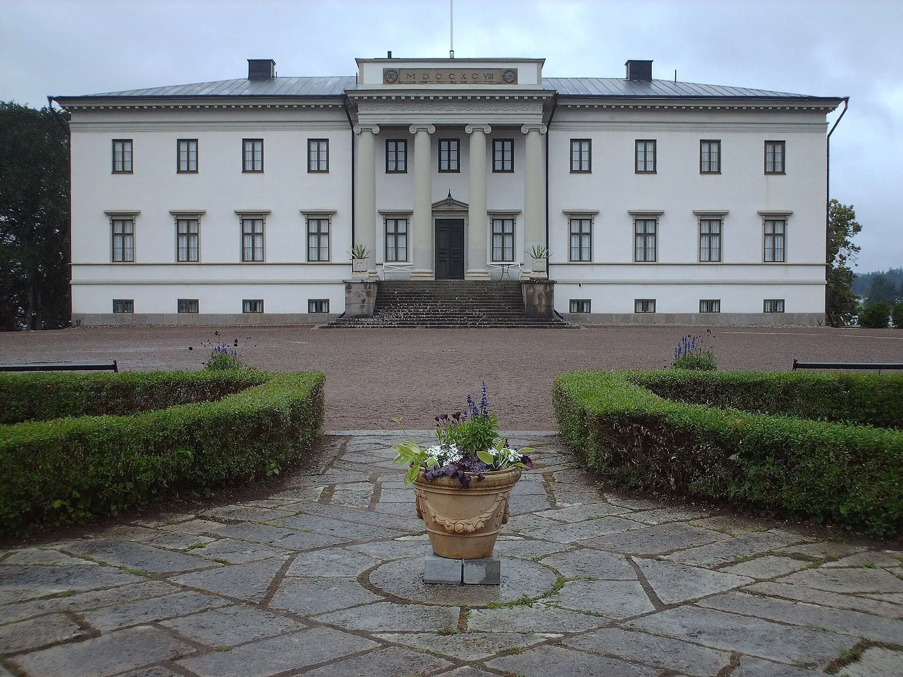 Stjernsunds slott sett framifrån: en ljus byggnad i två våningar med kolonner vid entrén, en stenlagd gård och klippta häckar.