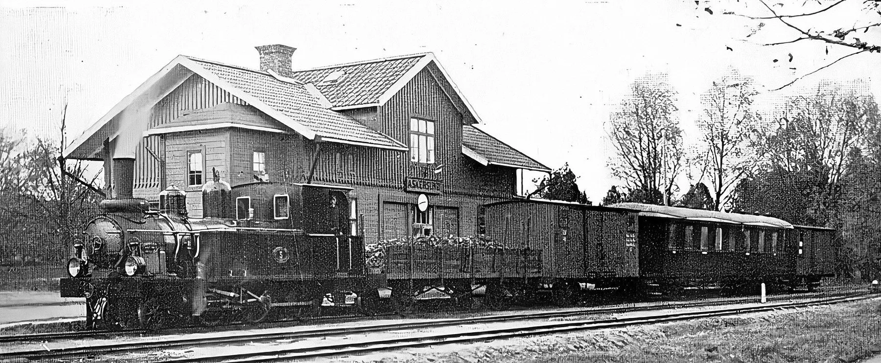 Svartvitt fotografi av ett ånglok med godsvagnar och personvagnar framför Askersunds stationshus i trä, 1926.