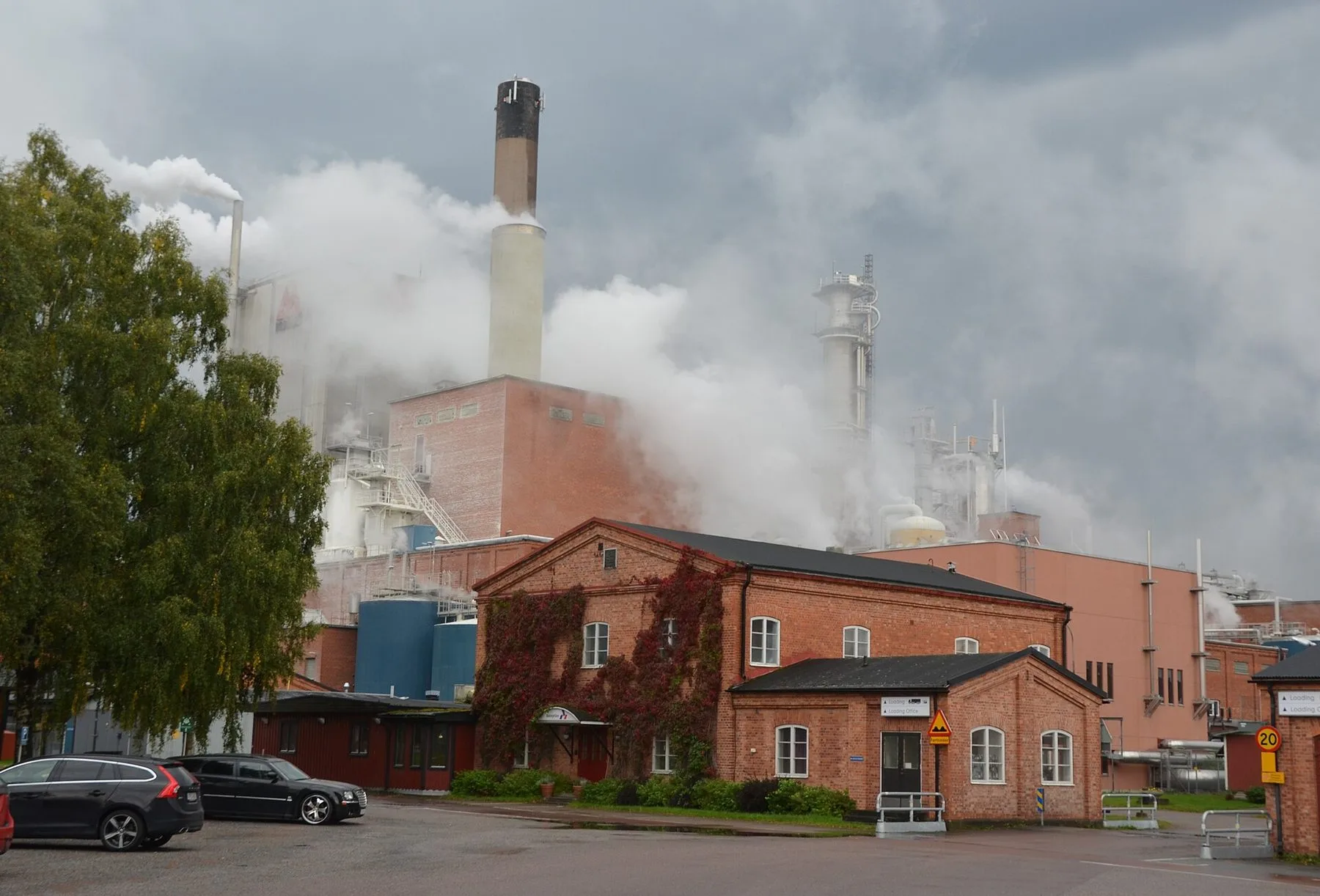 Massabruket Aspa bruk med en hög skorsten och stora ångmoln över fabriksbyggnader i rött tegel, och ett äldre tegelhus med vildvin framför en parkering.