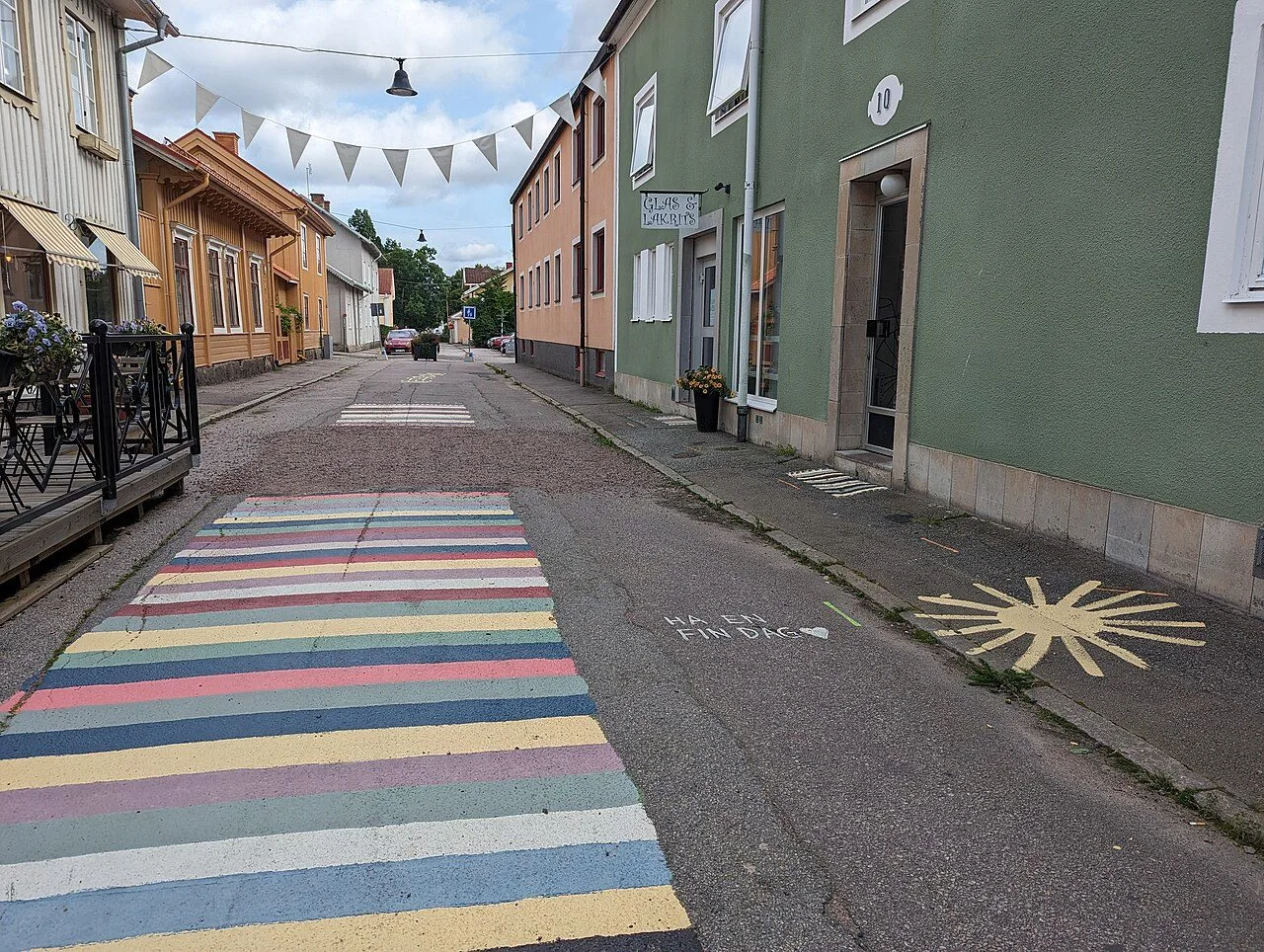En smal gata i Askersund med ett brett, regnbågsrandigt fält målat på asfalten, vimplar över gatan och trä- och stenhus med butiker.
